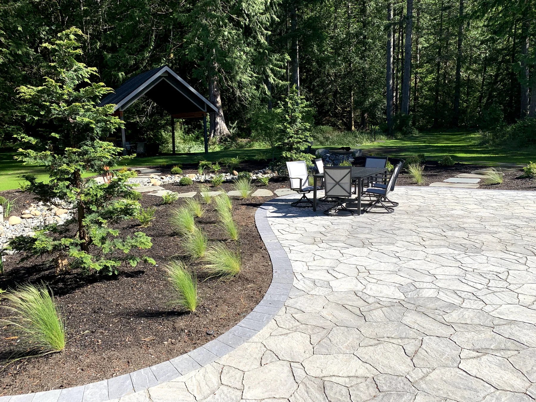 A generous patio is framed by a curved planting bed and woodland.
