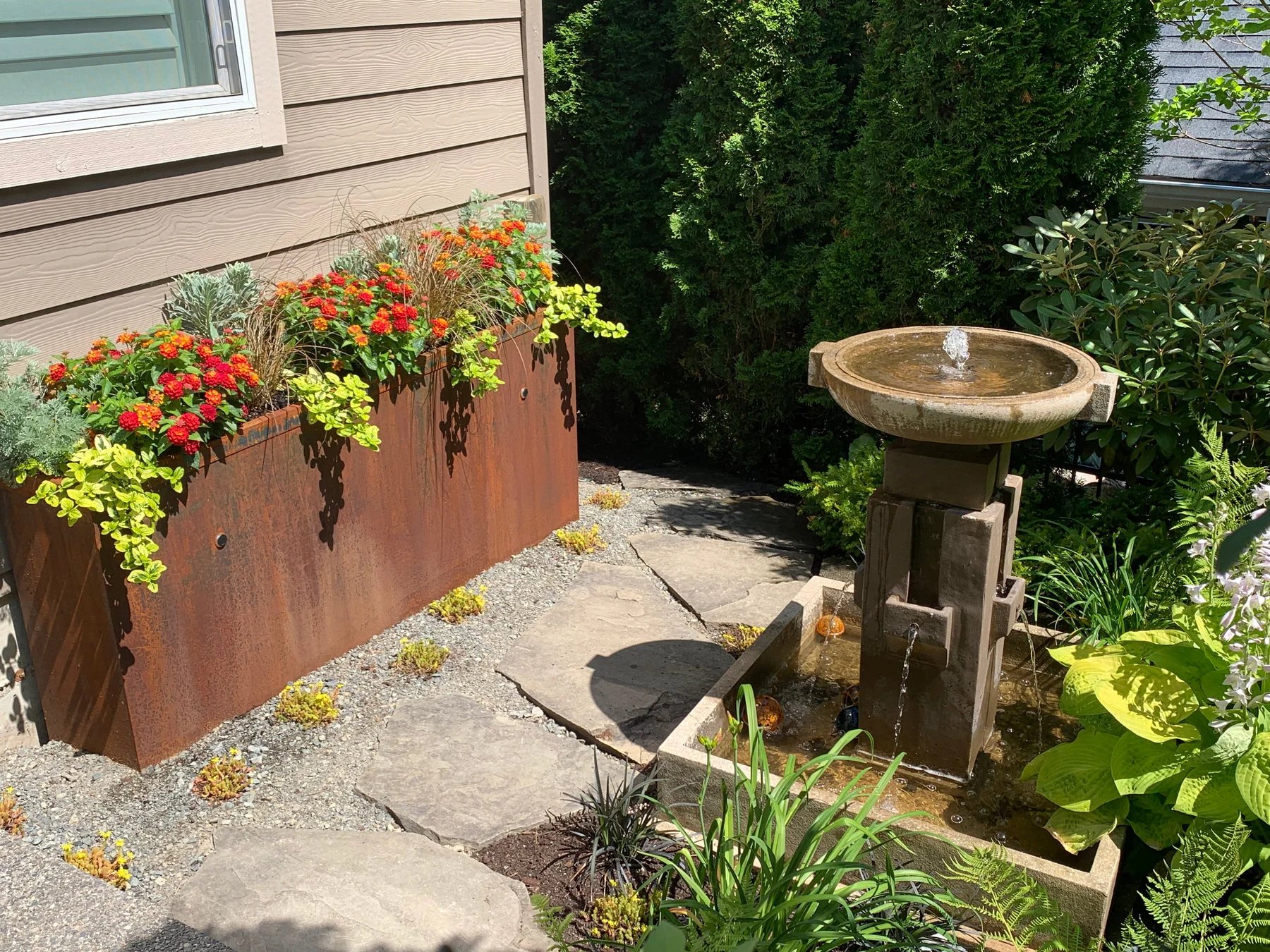 A Corten steel planter with colorful planting beside a stone birdbath.